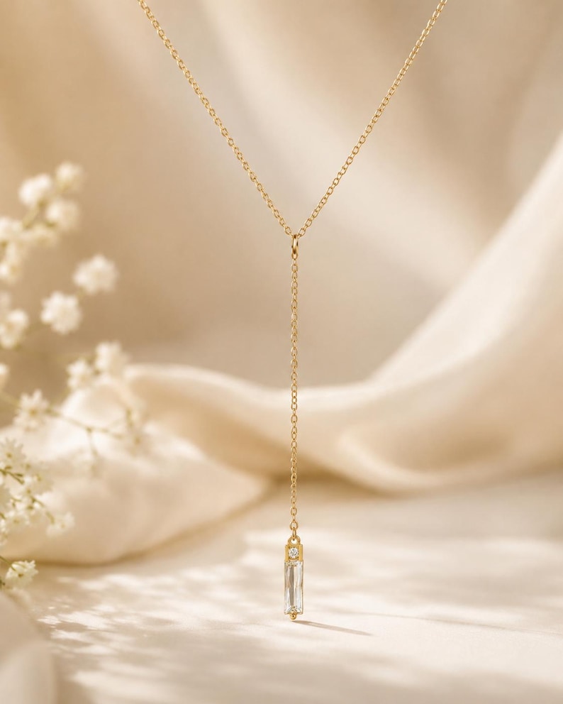 May include: A gold-toned Y-shaped necklace featuring a delicate chain and a vertical pendant. The pendant consists of a small, clear, rectangular baguette-cut stone topped with a tiny, round, clear accent stone, creating a minimalist drop design.