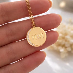 May include: Gold-colored necklace with a circular pendant. The pendant features the number "11:11" engraved in the center, with a crescent moon design encrusted with small, clear stones along the edge. The necklace is held by a person.
