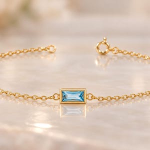 May include: A delicate gold chain bracelet featuring a single rectangular baguette-cut light blue gemstone set in a gold bezel. The bracelet includes a small circular spring ring clasp at one end and a matching jump ring at the other end.
