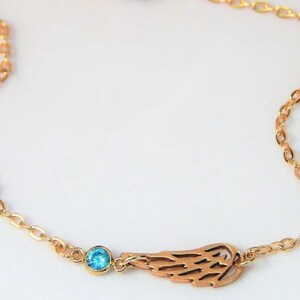 May include: A gold-colored necklace featuring a delicate chain. The necklace has a charm with a cut-out wing design and a small, round, light blue gemstone. The necklace is displayed against a white background.