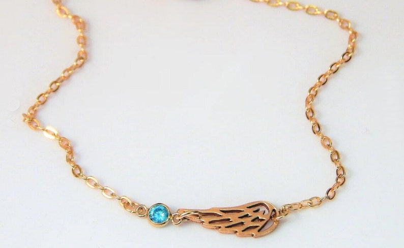 May include: A gold-colored necklace featuring a delicate chain. The necklace has a charm with a cut-out wing design and a small, round, light blue gemstone. The necklace is displayed against a white background.