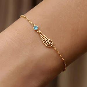 Puede incluir: Una delicada pulsera dorada con una pequeña gema azul claro y un dije en forma de ala. La pulsera está hecha de una fina cadena y está diseñada para ser usada en la muñeca.