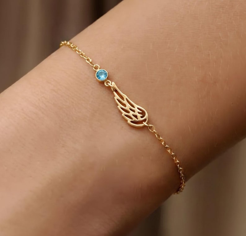 May include: A delicate gold-colored bracelet featuring a small, light blue gemstone and a wing-shaped charm. The bracelet is made of a fine chain and is designed to be worn on the wrist.