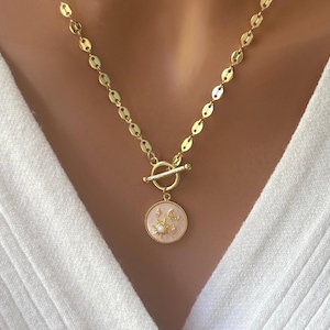 Op de afbeelding: Een gouden ketting met een toggle sluiting en een ronde hanger met een zon en maan ontwerp met kleine parel accenten.