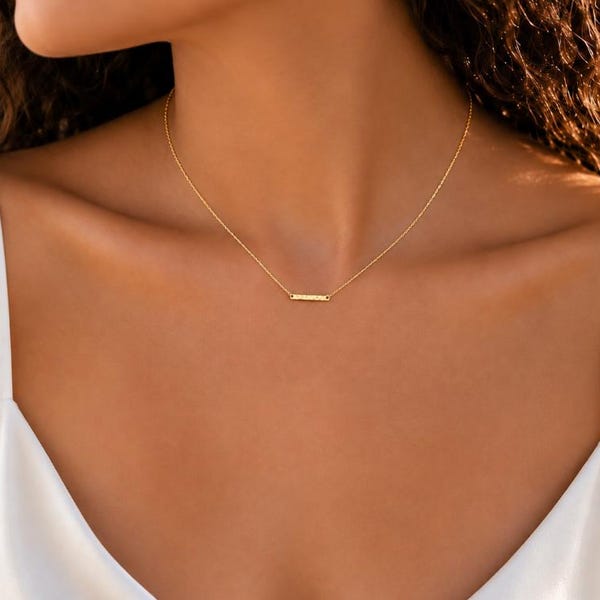 Könnte beinhalten: Eine zarte goldene Kette mit einem kleinen, horizontalen, rechteckigen Stabanhänger, der mittig an der Kette sitzt. Die Halskette wird um den Hals getragen, liegt im Bereich des Schlüsselbeins und wird mit einem weißen ärmellosen Oberteil kombiniert.