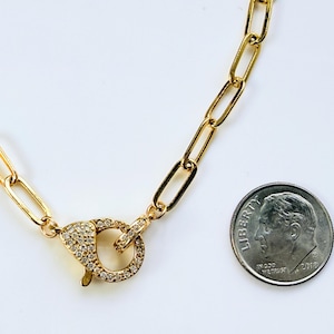 May include: Gold-colored chain necklace with a large clasp encrusted with small, clear gemstones. The chain links are rectangular. A U.S. dime is included for size reference.