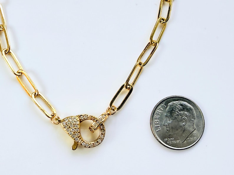 May include: Gold-colored chain necklace with a large clasp encrusted with small, clear gemstones. The chain links are rectangular. A U.S. dime is included for size reference.