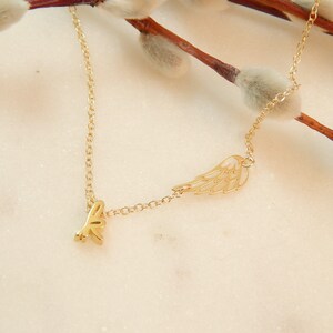 Puede incluir: Un collar de oro con un delicado dije de ala y un peque&ntilde;o dije de 'love'. El collar est&aacute; sobre un fondo blanco.