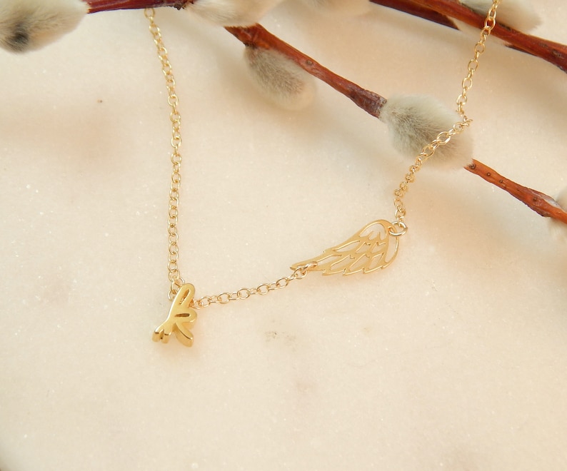 Puede incluir: Un collar de oro con un delicado dije de ala y un peque&ntilde;o dije de 'love'. El collar est&aacute; sobre un fondo blanco.