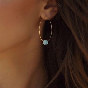 May include: Gold-tone hoop earrings with a small, light blue gemstone accent. The earrings are oval-shaped and appear to be lightweight. The gemstone is a pale blue color and adds a subtle pop of color to the design. These earrings are a simple yet elegant accessory.