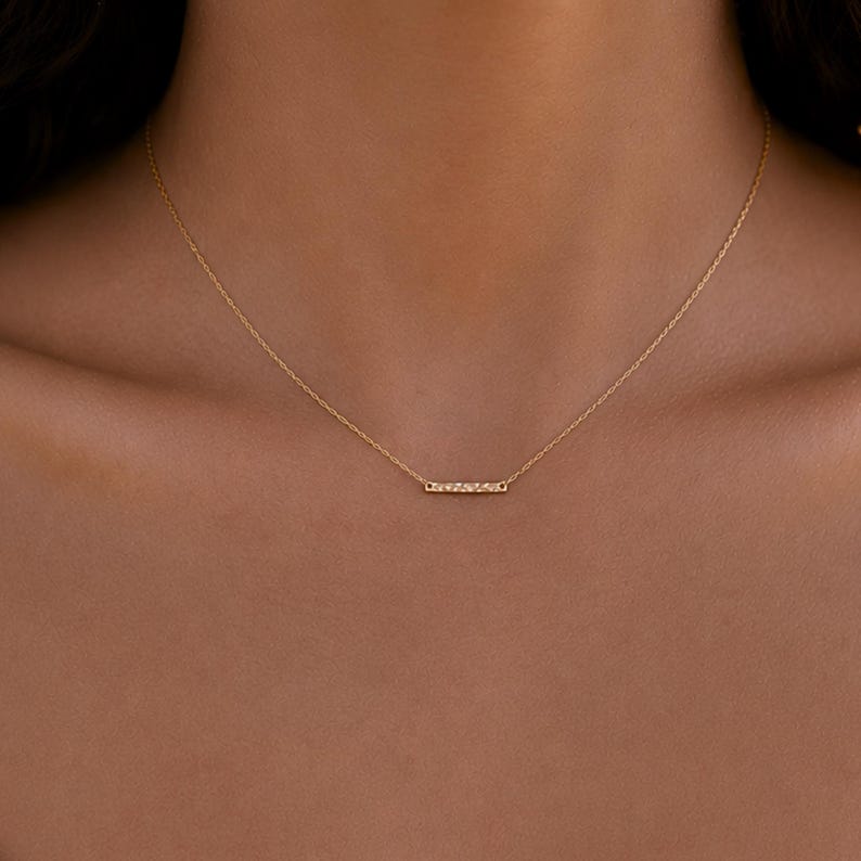 Puede incluir: Un delicado collar de cadena dorada que presenta un peque&ntilde;o colgante de barra rectangular horizontal con una superficie texturizada y brillante. La fina cadena dorada pulida se conecta a cada extremo de la barra, creando una joya sencilla.