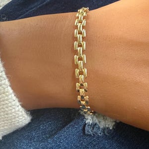 May include: A gold-colored bracelet with a rectangular link design. The bracelet is worn on a wrist, and the links are connected in a repeating pattern. The bracelet is made of a shiny, reflective material.