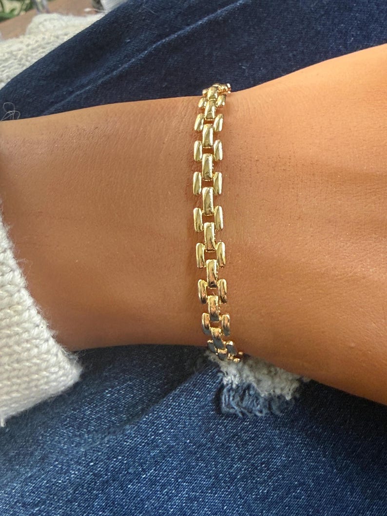 May include: A gold-colored bracelet with a rectangular link design. The bracelet is worn on a wrist, and the links are connected in a repeating pattern. The bracelet is made of a shiny, reflective material.