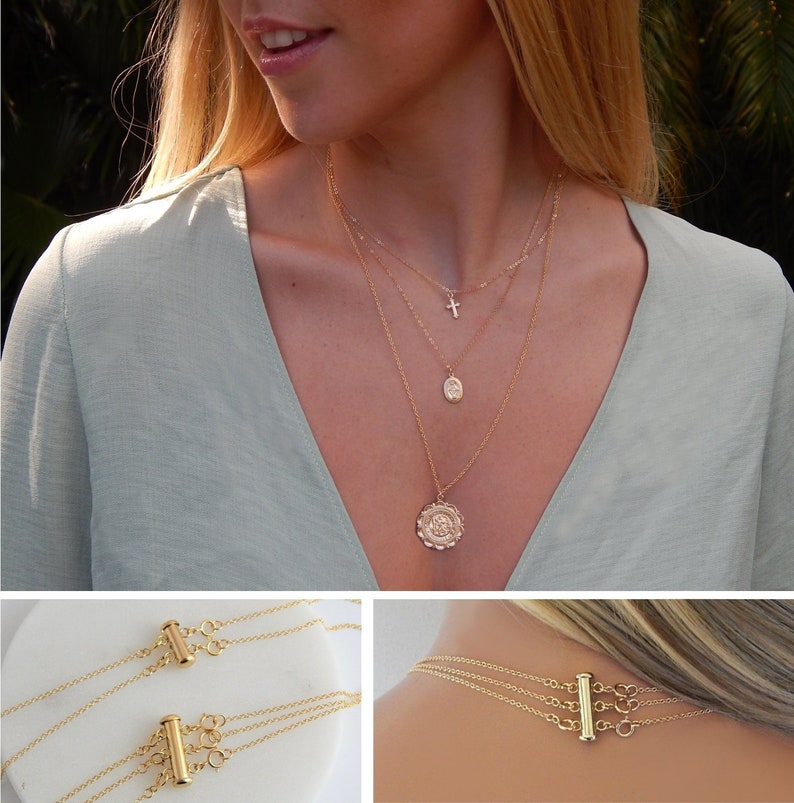 May include: A gold-colored layered necklace with three strands. The top strand features a small cross pendant, the middle a small oval pendant, and the bottom a round pendant with a floral design. The necklace is displayed on a person wearing a light green top.