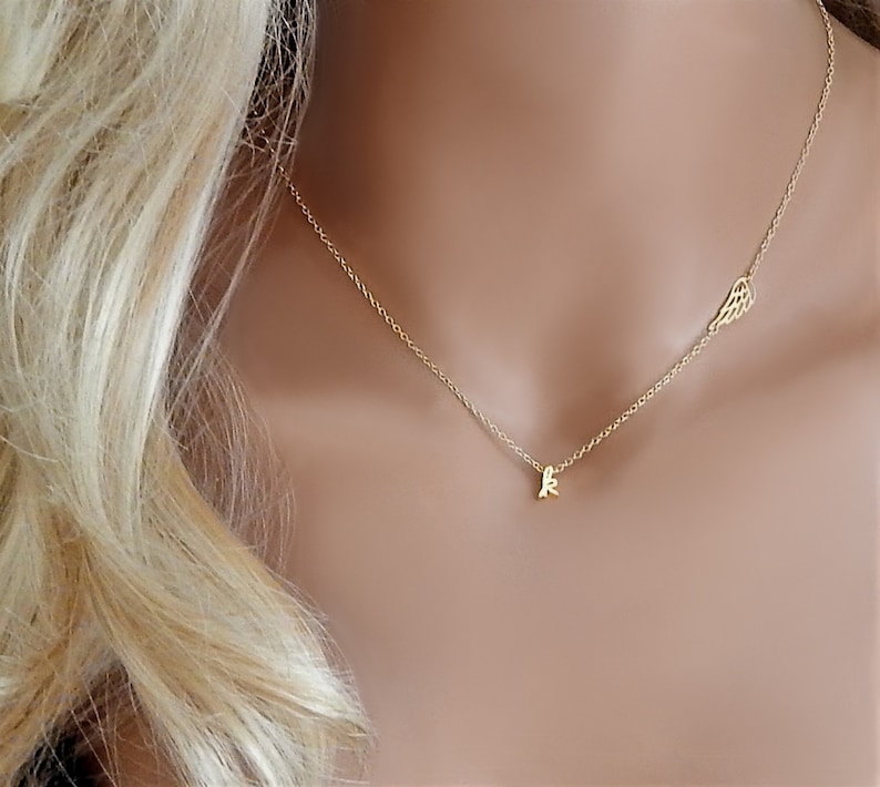 Puede incluir: Un collar de oro con una cadena delicada y un peque&ntilde;o dije con la letra 'K'. El collar tambi&eacute;n tiene un dije de ala en el otro lado.