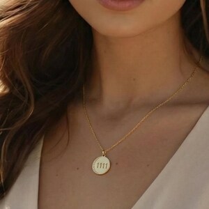 May include: A gold-colored necklace with a circular pendant. The pendant has the number "1111" engraved on it. The necklace is delicate and appears to be made of a thin chain. The pendant has a textured border.