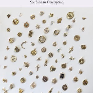 May include: A collection of gold-toned charms in various shapes and sizes, including suns, moons, crosses, and hearts. The charms are displayed on a white surface. Text at the top reads "For Add on Charms See Link in Description."