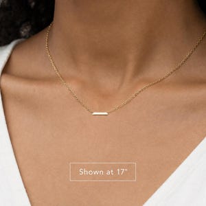 Puede incluir: Un collar de tono dorado que presenta una cadena delicada y un peque&ntilde;o colgante de barra rectangular horizontal. El collar se muestra con una longitud de 17 pulgadas, seg&uacute;n lo indicado por el texto en la imagen. El dise&ntilde;o es simple y refinado.