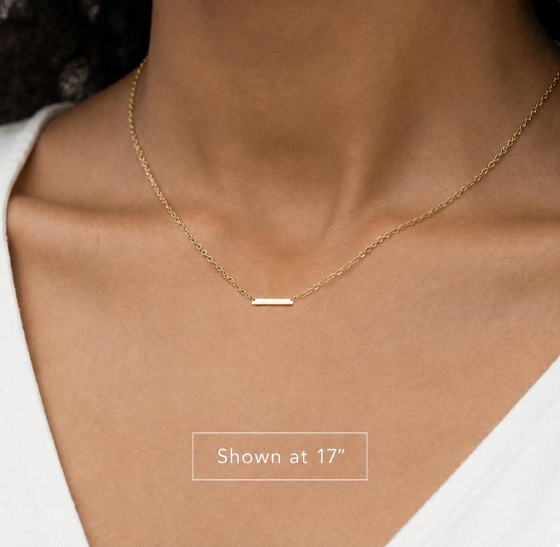 Puede incluir: Un collar de tono dorado que presenta una cadena delicada y un peque&ntilde;o colgante de barra rectangular horizontal. El collar se muestra con una longitud de 17 pulgadas, seg&uacute;n lo indicado por el texto en la imagen. El dise&ntilde;o es simple y refinado.