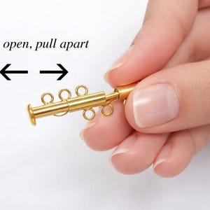 May include: A gold-colored jewelry clasp with multiple loops and a cylindrical end is held. The text "To open, pull apart" is above the clasp, with arrows indicating the direction to open. The clasp is designed to connect jewelry pieces.