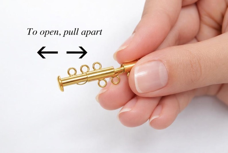 May include: A gold-colored jewelry clasp with multiple loops and a cylindrical end is held. The text "To open, pull apart" is above the clasp, with arrows indicating the direction to open. The clasp is designed to connect jewelry pieces.
