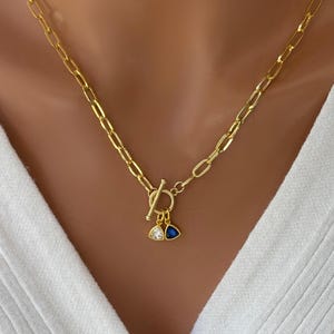 Op de afbeelding: Een gouden ketting met een toggle sluiting en twee hangende bedels. Een bedel is een gouden driehoek met een helder kristal, de andere is een gouden driehoek met een blauw kristal.