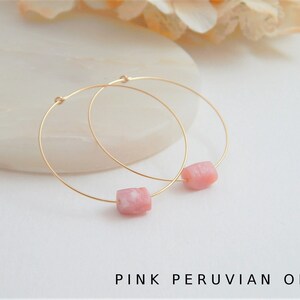 May include: A pair of gold hoop earrings, each featuring a pink Peruvian opal bead. The earrings are displayed on a white surface, with the text "PINK PERUVIAN OPAL" at the bottom. The hoops are delicate and the opals have a soft, natural color.