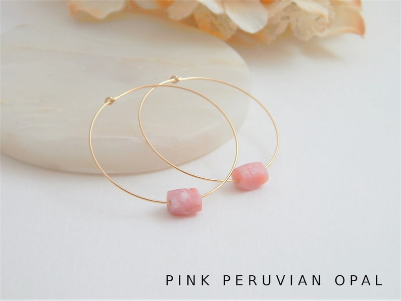 May include: A pair of gold hoop earrings, each featuring a pink Peruvian opal bead. The earrings are displayed on a white surface, with the text "PINK PERUVIAN OPAL" at the bottom. The hoops are delicate and the opals have a soft, natural color.