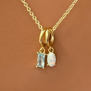 May include: Gold-toned necklace with two pendant charms. One charm is a rectangular, light blue gemstone, and the other is an oval, iridescent white gemstone. The chain and charms are set against a warm brown background.