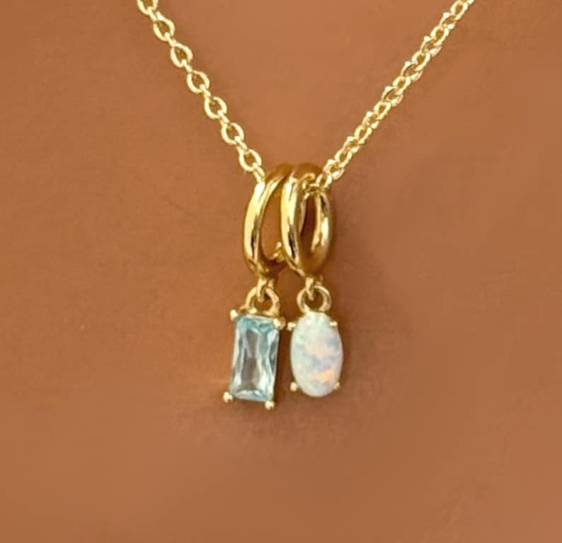 May include: Gold-toned necklace with two pendant charms. One charm is a rectangular, light blue gemstone, and the other is an oval, iridescent white gemstone. The chain and charms are set against a warm brown background.