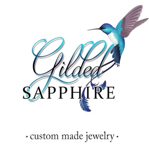 May include: The Gilded Sapphire logo features a hummingbird in shades of blue and purple, with the words "Gilded Sapphire" in elegant script. Below, the text reads "custom made jewelry."
