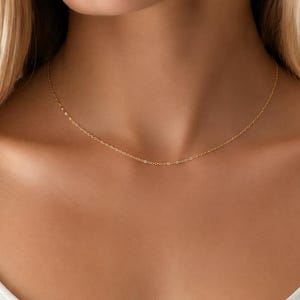 May include: A delicate gold chain necklace featuring a fine, thin link design that rests against the collarbone. The necklace is crafted from a polished yellow gold material, showcasing a simple, minimalist aesthetic with a secure clasp closure.