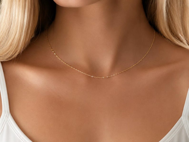 May include: A delicate gold chain necklace featuring a fine, thin link design that rests against the collarbone. The necklace is crafted from a polished yellow gold material, showcasing a simple, minimalist aesthetic with a secure clasp closure.