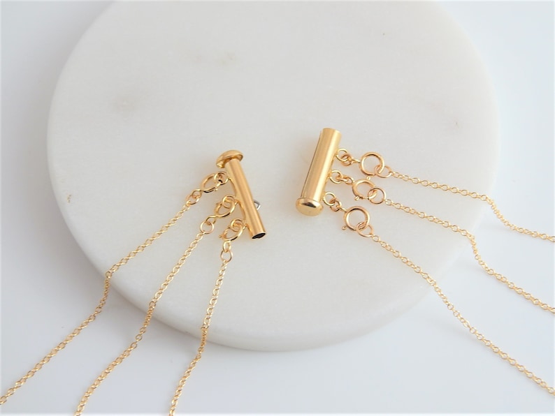 May include: Two gold-colored necklace clasps with multiple chain attachments. Each clasp has a cylindrical bar with three small loops for attaching chains. The chains are delicate and dangle from the clasps. The clasps are on a white surface.