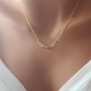 K&ouml;nnte beinhalten: Eine goldene Kettenkette mit einem zarten Paar Engelsfl&uuml;geln und einem kleinen blauen Edelstein in der Mitte.