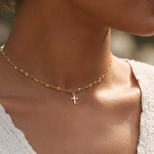 May include: A gold-toned necklace featuring a delicate chain with elongated, textured oval links and a small, simple cross pendant suspended from the center. The necklace rests against the neck, showcasing the metallic finish and the cross design.