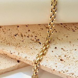 May include: A gold-colored bracelet with a repeating rectangular link design. The bracelet is displayed on a textured, light-colored surface, creating a contrast with the shiny metal. The links are closely connected, forming a continuous chain.