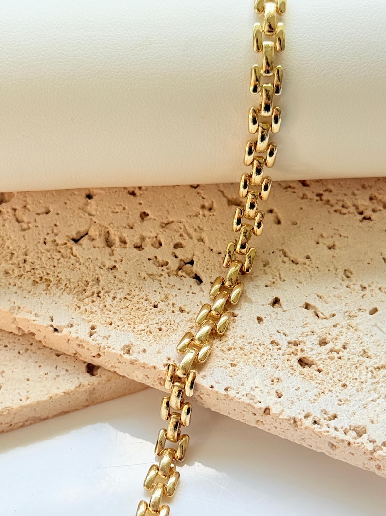 May include: A gold-colored bracelet with a repeating rectangular link design. The bracelet is displayed on a textured, light-colored surface, creating a contrast with the shiny metal. The links are closely connected, forming a continuous chain.