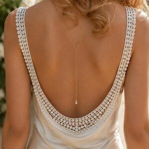May include: A cream-colored satin dress featuring a deep scoop back with wide straps adorned with a circular lace trim. A delicate gold-toned chain necklace hangs down the center of the back, ending in a single clear teardrop-shaped pendant.