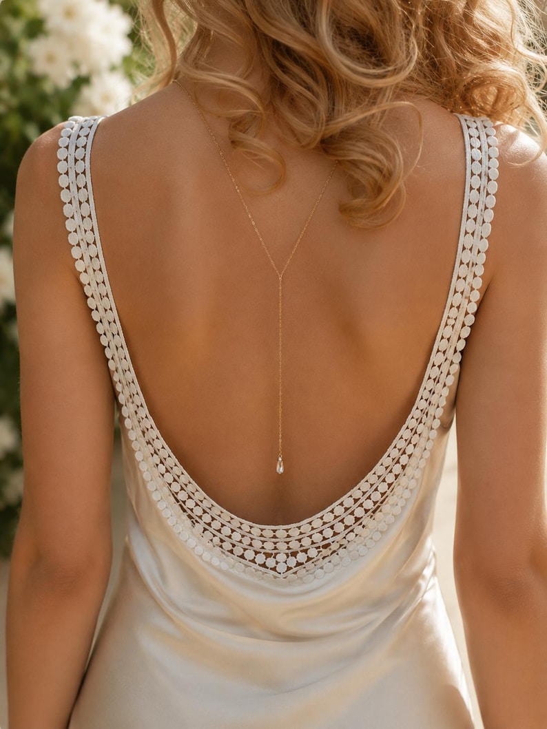 May include: A cream-colored satin dress featuring a deep scoop back with wide straps adorned with a circular lace trim. A delicate gold-toned chain necklace hangs down the center of the back, ending in a single clear teardrop-shaped pendant.