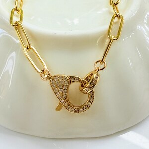 May include: Gold-colored necklace with a large clasp. The necklace features a chain of rectangular links. The clasp is encrusted with small, clear gemstones. The jewelry is displayed on a white surface.