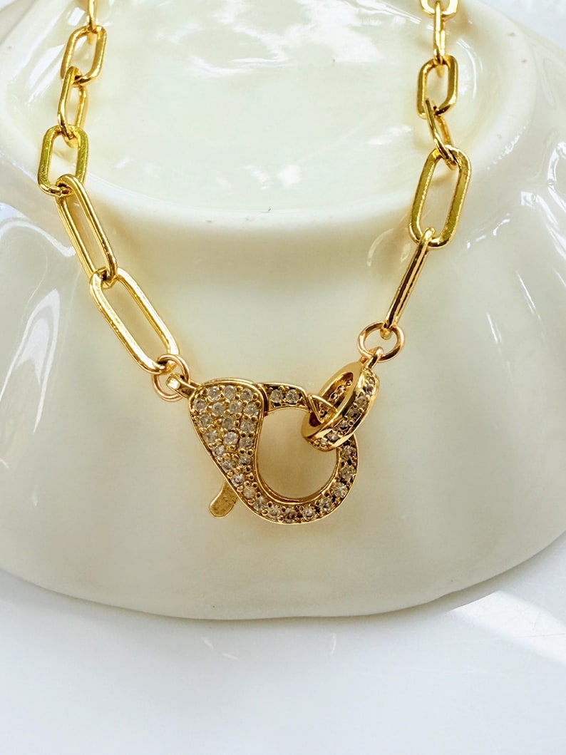 May include: Gold-colored necklace with a large clasp. The necklace features a chain of rectangular links. The clasp is encrusted with small, clear gemstones. The jewelry is displayed on a white surface.