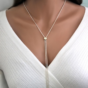 K&ouml;nnte beinhalten: Eine silberne Lariat-Kette mit einem kleinen runden Anh&auml;nger, der einen klaren Stein in einer goldenen Fassung tr&auml;gt. Die Kette besteht aus winzigen Perlen und hat einen zarten Look.