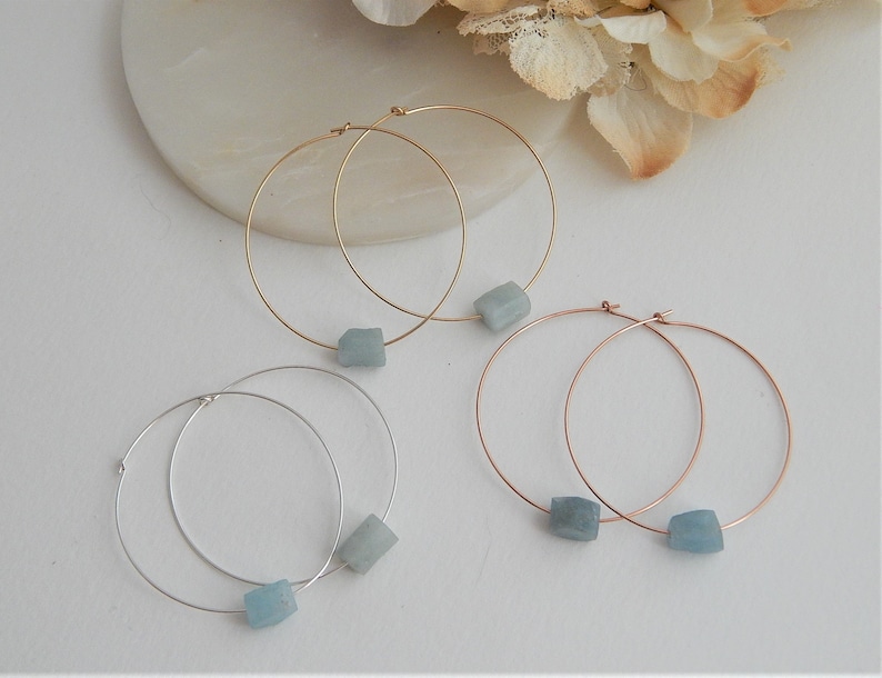 May include: Four pairs of hoop earrings in gold, silver, and rose gold tones. Each earring features a small, light blue, square-shaped gemstone. The earrings are displayed on a white surface.