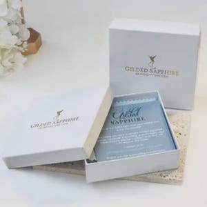 May include: White gift boxes with gold text and a hummingbird logo. One box is open, revealing a blue card with the text "Gilded Sapphire." The boxes are on a light-colored surface.