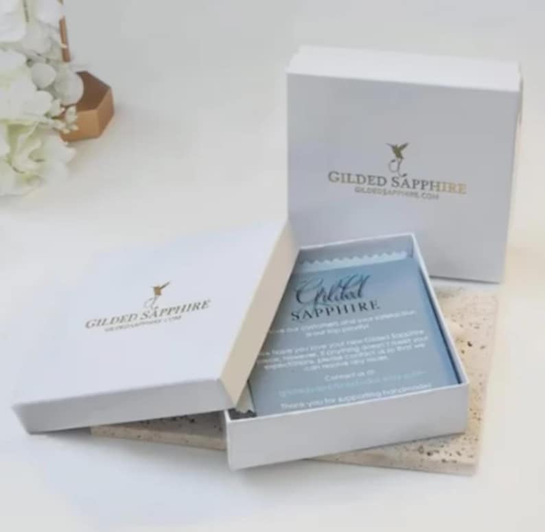 May include: White gift boxes with gold text and a hummingbird logo. One box is open, revealing a blue card with the text "Gilded Sapphire." The boxes are on a light-colored surface.