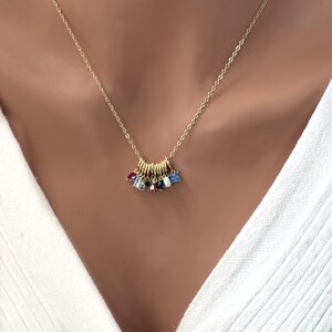 May include: A gold-colored necklace with a delicate chain. The pendant features a series of small gold rings, each adorned with a colorful gemstone. The gemstones include red, blue, green, and white stones. The necklace is worn against a white top.