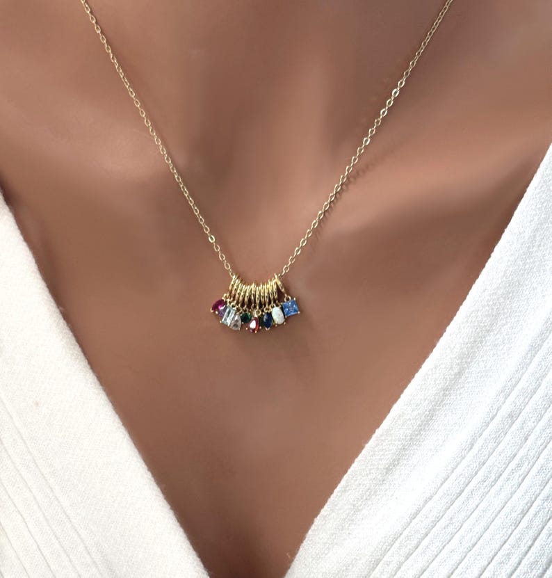 May include: A gold-colored necklace with a delicate chain. The pendant features a series of small gold rings, each adorned with a colorful gemstone. The gemstones include red, blue, green, and white stones. The necklace is worn against a white top.