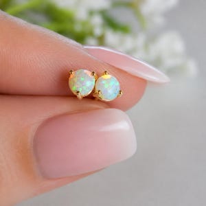 May include: A pair of round opal stud earrings set in gold metal prongs. Each earring features a circular opal gemstone displaying iridescent flashes of green, blue, and orange light, securely held by a three-prong gold setting.