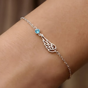 May include: A delicate silver bracelet featuring a small, round, light blue gemstone and a stylized wing charm. The bracelet is made of a fine chain and is designed to be worn on the wrist. The wing charm has intricate cut-out details.